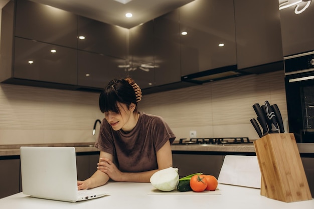 Heureuse belle jeune femme appréciant la cuisine à la recherche d'une recette sur l'ordinateur portable dans la cuisine