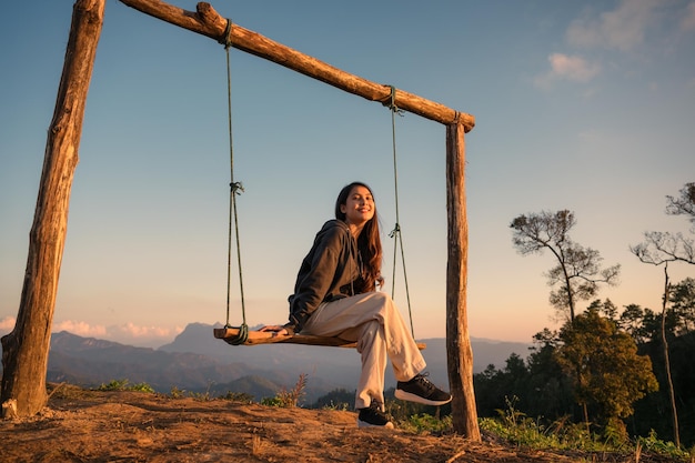 Heureuse belle jeune femme asiatique assise et souriante à la balançoire en bois au sommet de la montagne dans la campagne le soir