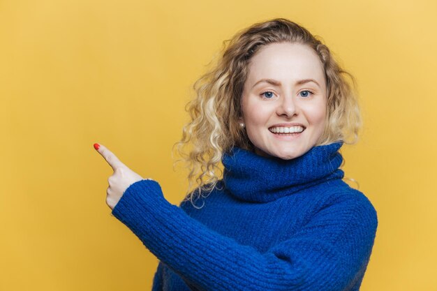 Heureuse belle jeune femme blonde a une expression heureuse porte un pull bleu décontracté indique à l'espace de copie vierge sur le mur de studio blanc jaune annonce quelque chose avec grand plaisir