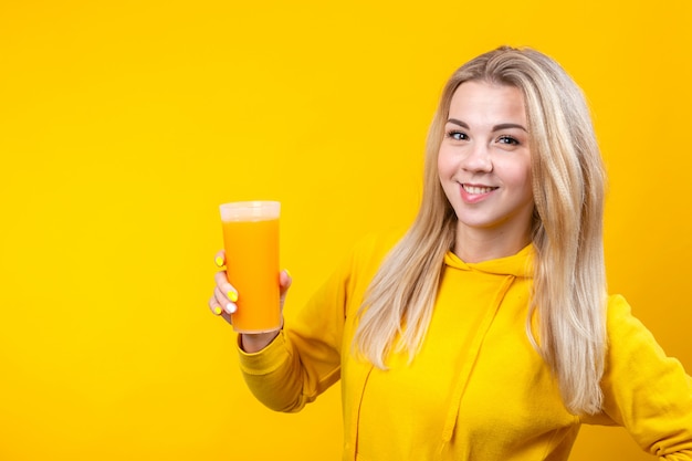 Heureuse belle jeune femme blonde en vêtements de sport décontractés jaunes tenant un verre de jus d'orange,