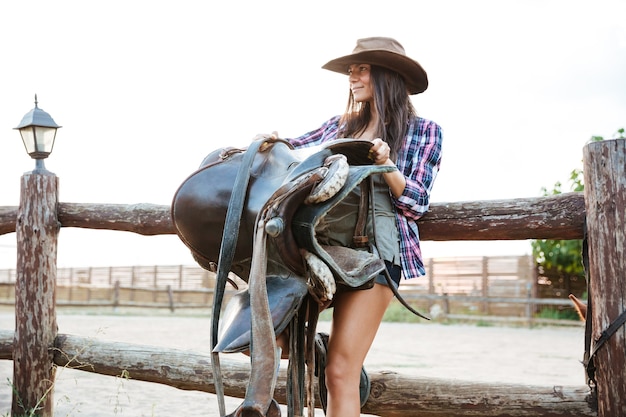 Heureuse belle jeune femme cow-girl au chapeau debout et tenant la selle pour monter à cheval