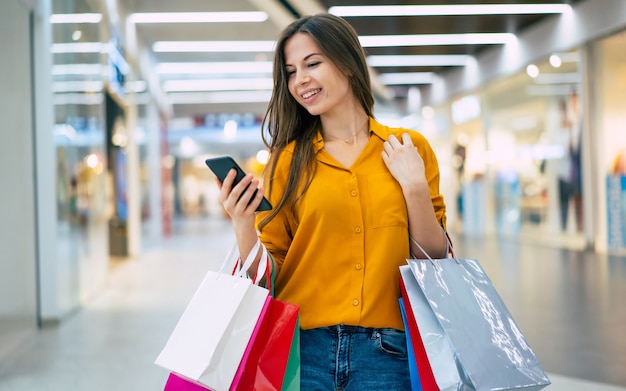 Heureuse belle jeune femme élégante avec des sacs à provisions utilise un téléphone intelligent tout en marchant dans le centre commercial