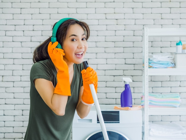 Heureuse belle jeune femme, femme au foyer portant des gants en caoutchouc orange, écouter de la musique avec un casque vert chantant amusant avec une vadrouille comme un microphone sur un mur de briques blanches à la maison.