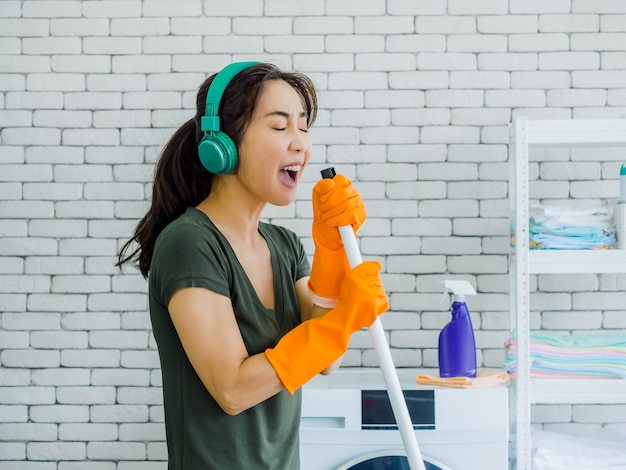 Heureuse belle jeune femme, femme au foyer portant des gants en caoutchouc orange, écouter de la musique avec un casque vert chantant amusant avec une vadrouille comme un microphone sur un mur de briques blanches à la maison.