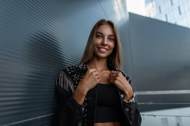 Heureuse belle jeune femme avec un joli sourire dans une veste en jean élégante à la mode dans un T-shirt noir se tient près d'un bâtiment moderne en métal - verre dans la ville