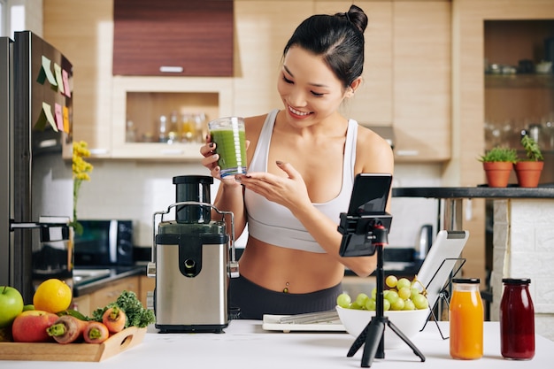 Heureuse belle jeune femme montrant un verre de smoothie vert qu'elle a fait à ses abonnés lors du tournage du blog