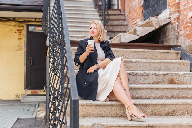 Heureuse belle jeune femme en robe et veste se promène dans la ville et boit du café