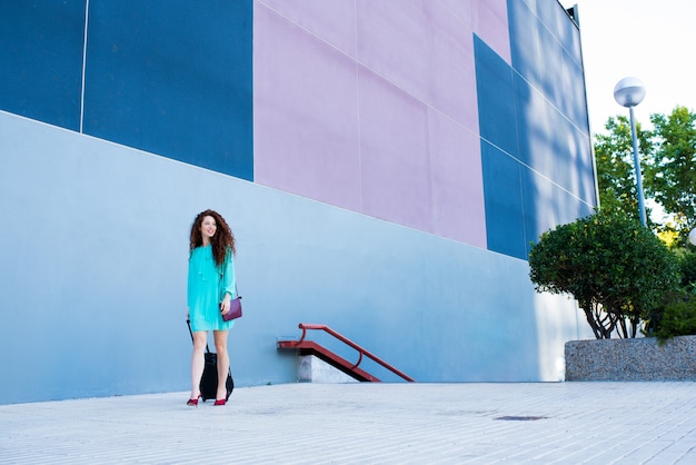 Heureuse belle jeune femme rousse avec une valise marchant sur un mur bleu