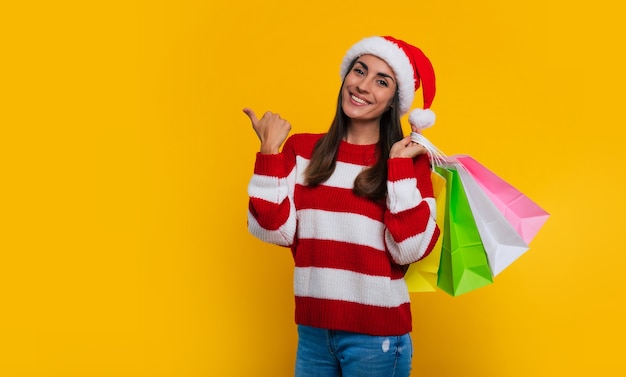 Heureuse belle jeune femme souriante dans un bonnet de Noel de Noël avec des sacs à provisions colorés dans ses mains pointe vers un fond jaune