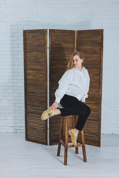 Heureuse belle jeune femme avec un sourire dans une tenue à la mode avec une chemise blanche en cuir des chaussures beiges et un pantalon noir est assise sur une chaise en bois