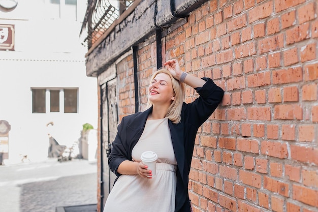 Heureuse belle jeune femme vêtue d'une robe et d'une veste se tient près d'un mur de briques et boit du café