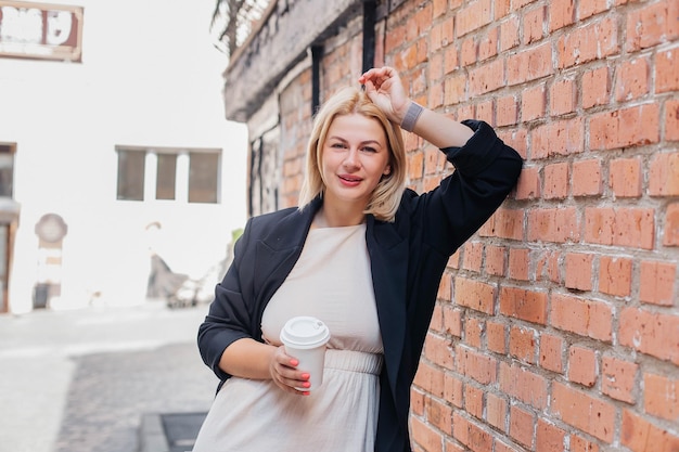 Heureuse belle jeune femme vêtue d'une robe et d'une veste se tient près d'un mur de briques et boit du café