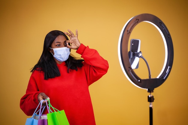 Heureuse blogueuse afro-américaine dans un masque de protection contre les virus avec une lampe annulaire de sacs multicolores