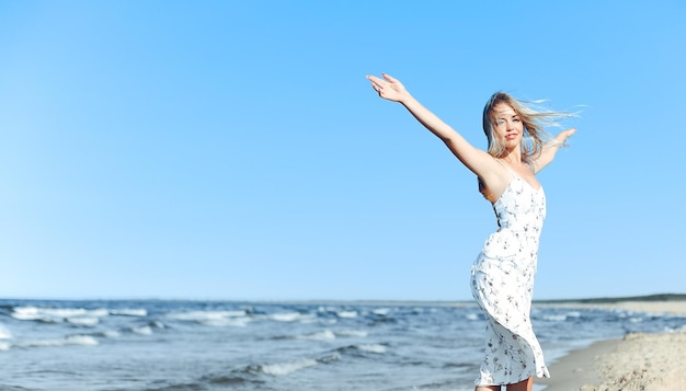 Heureuse blonde belle femme sur la plage de l'océan debout dans une robe d'été blanche, bras ouverts.