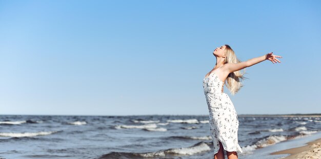 Heureuse blonde belle femme sur la plage de l'océan debout dans une robe d'été blanche, bras ouverts.