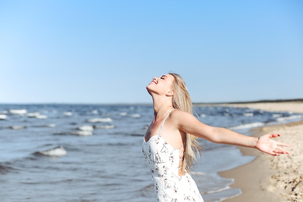 Heureuse blonde belle femme sur la plage de l'océan debout dans une robe d'été blanche, levant les mains.