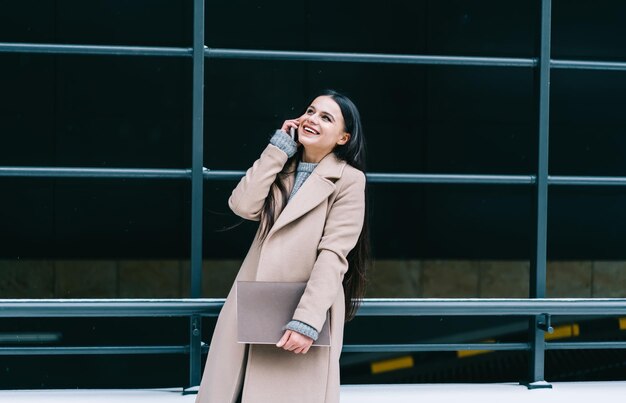 Heureuse brune en manteau beige tenant un bloc-notes et riant tout en parlant au téléphone portable debout