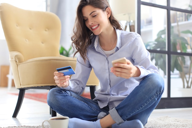 Heureuse brune naturelle utilisant une carte de crédit et un téléphone portable dans la chambre.