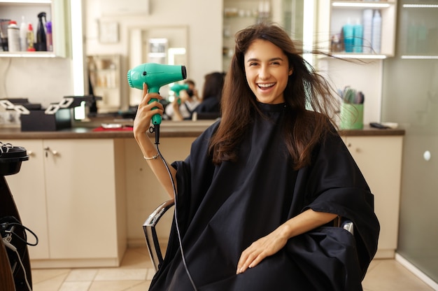 Heureuse cliente à l'aide de sèche-cheveux dans un salon de coiffure. Femme assise sur une chaise dans un salon de coiffure. Entreprise de beauté et de mode, service professionnel