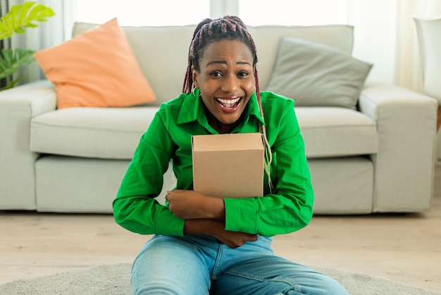 Heureuse dame afro-américaine étreignant un colis en carton se faisant livrer une commande en ligne et souriant à la caméra