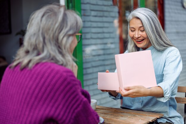 Heureuse dame asiatique âgée ouvre un cadeau avec son meilleur ami célébrant des vacances au café