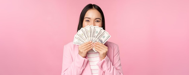 Heureuse dame asiatique en costume tenant de l'argent dollars avec une expression de visage heureux debout sur fond rose
