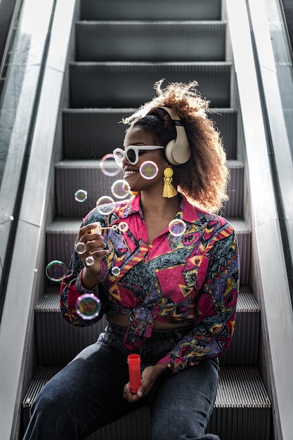 Photo heureuse dame noire dans les écouteurs faisant des bulles sur l'escalator