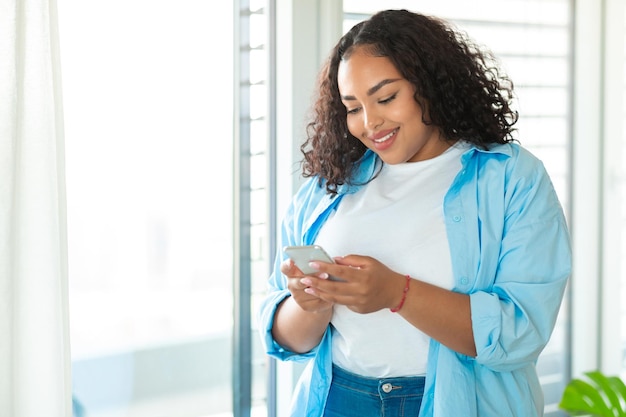 Heureuse dame obèse noire utilisant un téléphone portable et envoyant des SMS debout près de la fenêtre à l'espace de copie à la maison
