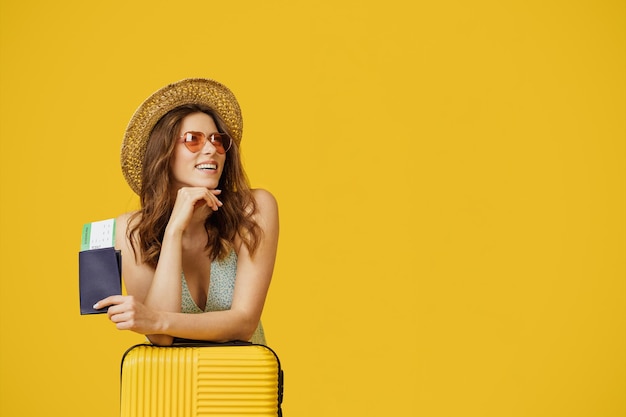 Heureuse dame avec passeport et billets d'avion s'appuyant sur une valise et regardant de côté l'espace libre sur fond jaune