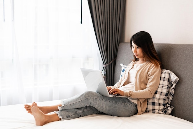 Heureuse et décontractée belle femme asiatique travaillant sur un ordinateur portable assis sur le lit dans la maison La technologie et le style de vie travaillent à partir du concept Copier l'espace