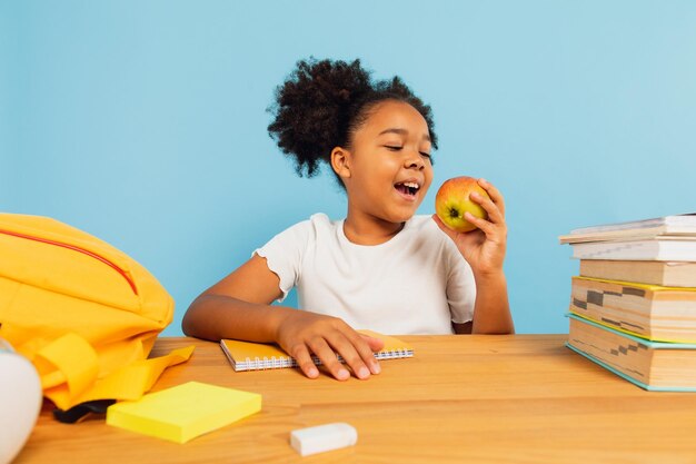 Heureuse écolière afro-américaine assise au bureau dans la salle de classe et déjeunant avec pomme sur fond bleu Concept de retour à l'école