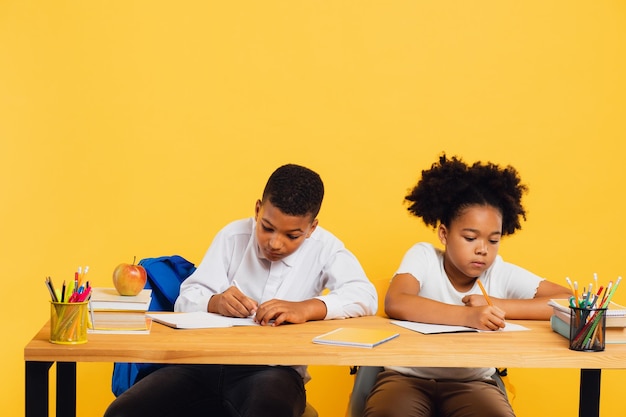 Heureuse écolière afro-américaine et écolier métis assis ensemble au bureau et étudiant sur fond jaune Concept de retour à l'école