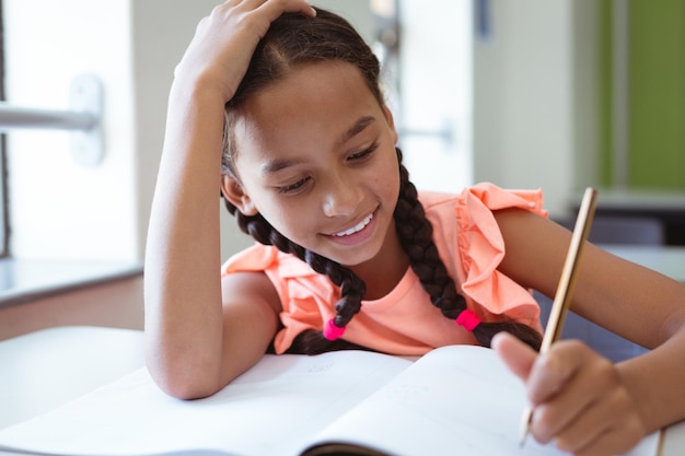 Heureuse écolière métisse en classe assise au bureau souriant et écrivant dans un livre