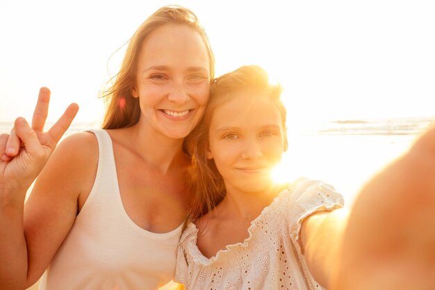 Heureuse élégamment mère et fille prenant selfie sur une plage de sable au coucher du soleilmères jourpetite fille blonde et belle femme prenant des photos au téléphone tourisme à l'étranger appel vidéo en ligne