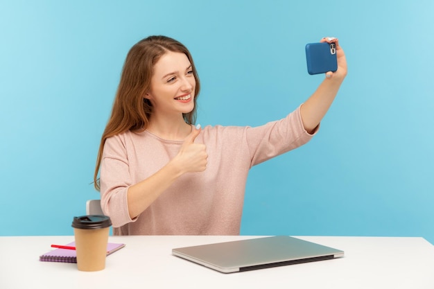 Heureuse employée de bureau ou blogueuse satisfaite assise sur le lieu de travail et prenant un selfie par téléphone portable parlant par appel vidéo avec le pouce levé comme un geste en studio intérieur tourné sur fond bleu