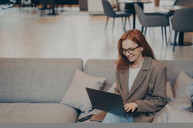 Heureuse employée naviguant sur le Web alors qu'elle était assise sur un canapé dans une salle de pause dans un bureau moderne