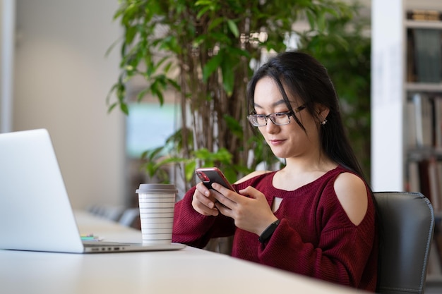 Heureuse étudiante asiatique distraite de l'étude à l'aide d'un smartphone alors qu'elle était assise dans la bibliothèque