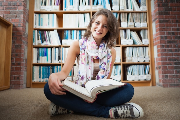 Heureuse étudiante contre bibliothèque avec un livre sur le sol de la bibliothèque