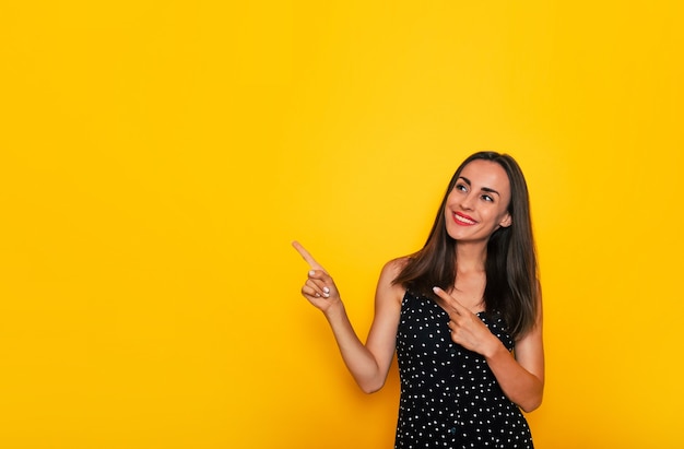 Heureuse excitée magnifique femme brune souriante dans une robe d'été noire pose isolée sur fond jaune et pointant vers l'extérieur