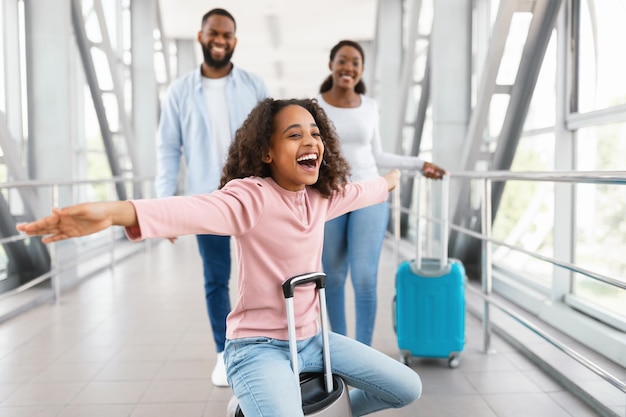 Heureuse famille aimante à l'aéroport. Portrait d'une joyeuse fille noire excitée assise sur une valise et riant, écartant les bras imitant l'avion, souriant des parents joyeux marchant sur fond flou
