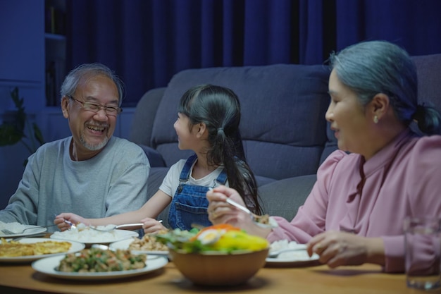 Heureuse famille asiatique, grand-mère, grand-père et petite-fille, dînant sur une table et s'amusant pendant la nuit à la maison, un parent âgé et un enfant mangeant de la nourriture ensemble dans le salon à l'intérieur