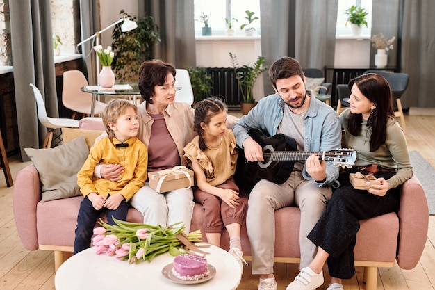 Heureuse famille de cinq personnes assises en rang sur un grand canapé confortable et moelleux dans le salon, parlant et écoutant le chant d'un jeune homme à la guitare