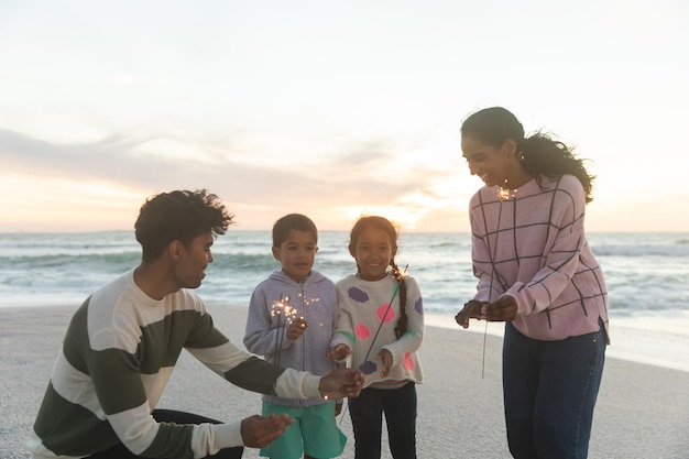 Heureuse famille multiraciale appréciant avec des cierges magiques à la plage contre le ciel au coucher du soleil