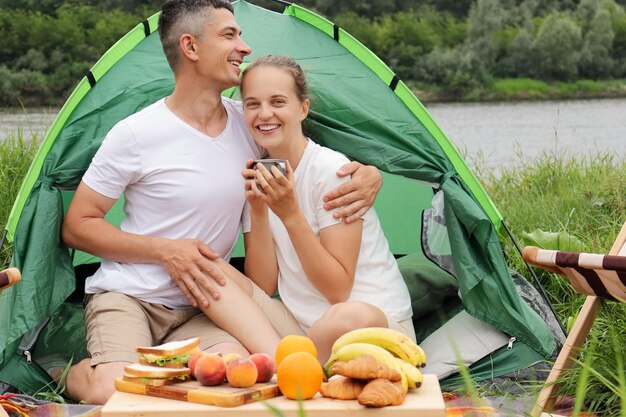 Heureuse famille souriante et aimante se reposant en plein air Femme et homme romantique portant des vêtements décontractés assis à la tente près de la rivière buvant du café ou du thé s'étreignant en profitant du temps ensemble