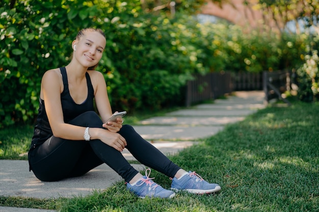 Heureuse femme adulte aux cheveux noirs en vêtements de sport utilise un smartphone pour bloguer et discuter en ligne satisfaite des résultats d'entraînement pose sur fond de nature a une pause après l'entraînement de fitness