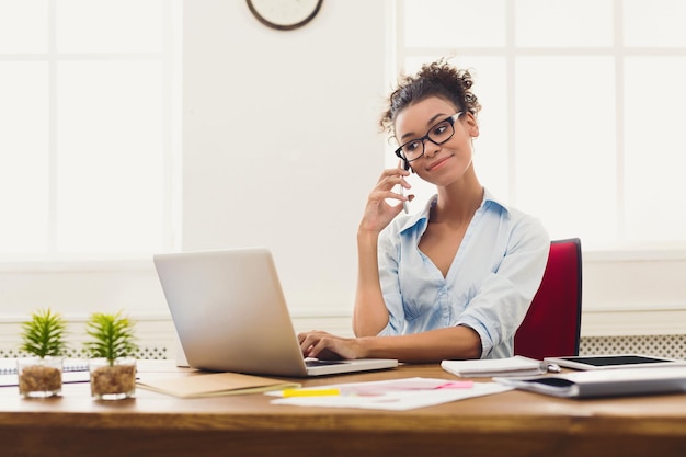 Heureuse femme d'affaires afro-américaine parlant sur mobile au bureau. Jeune femme gestionnaire consultant au téléphone, utilisant un ordinateur portable sur le lieu de travail, espace de copie