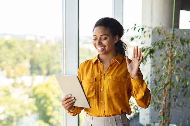 Heureuse femme d'affaires afro-américaine regardant la tablette et agitant la main à la webcamera travaillant à l'intérieur du bureau