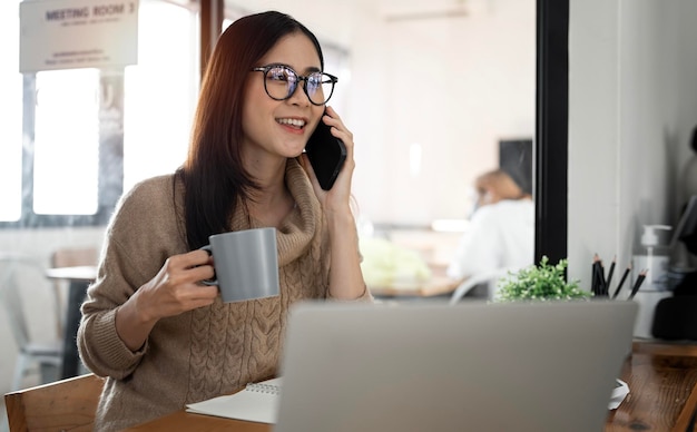 Heureuse femme d'affaires assise au bureau derrière son ordinateur portable et parlant avec quelqu'un sur son téléphone portable tout en travaillant dans un espace de travail