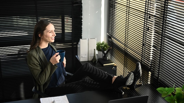 Heureuse femme d'affaires caucasienne se reposant sur une chaise de bureau confortable et buvant du café