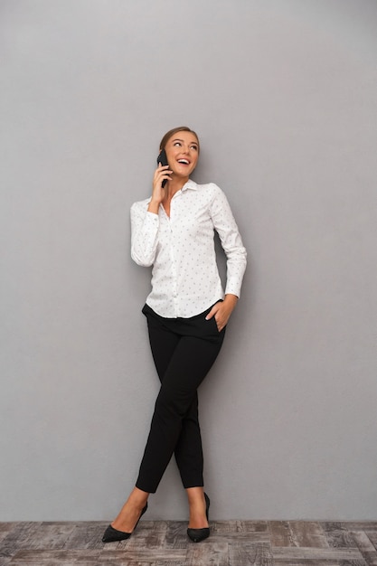 Heureuse femme d'affaires debout sur fond de mur gris parlant par téléphone mobile.